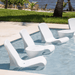 four white pool chairs and two white pool side table in a pool beside grassy field.