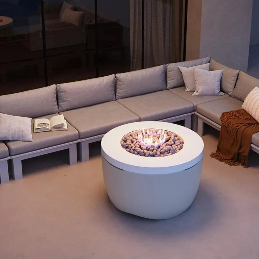 White round hidden propane tank fire pit placed on a patio surrounded with grey sectional sofa beside glass wall