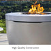 Light grey round hidden propane tank fire pit with lava rocks on fire on top placed beside metal fence