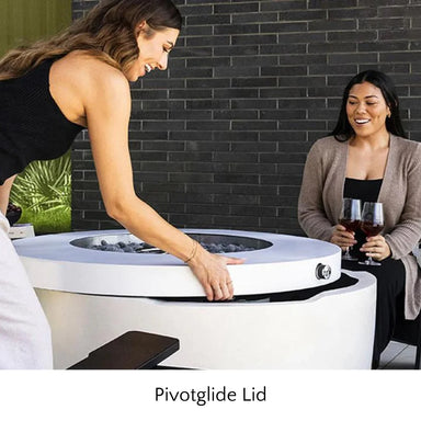 A woman opening the top of white round hidden propane tank fire pit while another woman sitting beside holding two glasses of wine