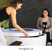 A woman opening the top of white round hidden propane tank fire pit while another woman sitting beside holding two glasses of wine