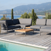 A square wooden outdoor coffee table with gray aluminum frame set on a wooden deck poolside with dark gray sectional sofa