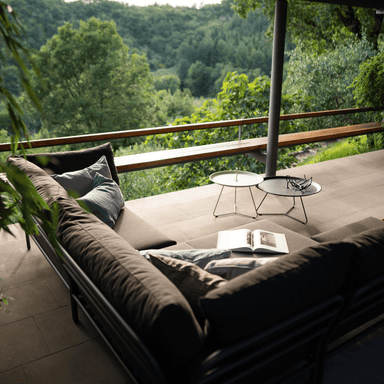 A brown outdoor sectional sofa with 2 outdoor tray table set on a tiled patio surrounded by greenery