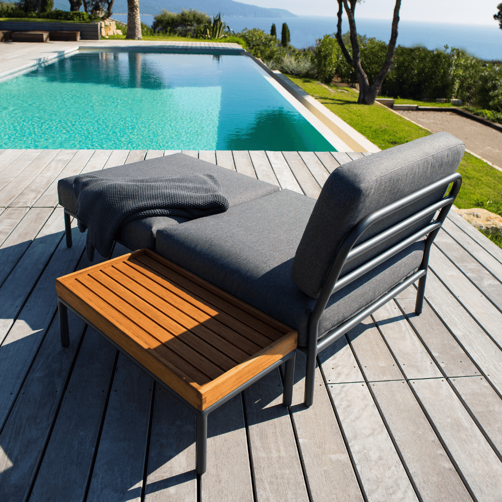 A dark gray outdoor lounge chair and dark gray outdoor ottoman with rectangular wooden outdoor side table placed at poolside