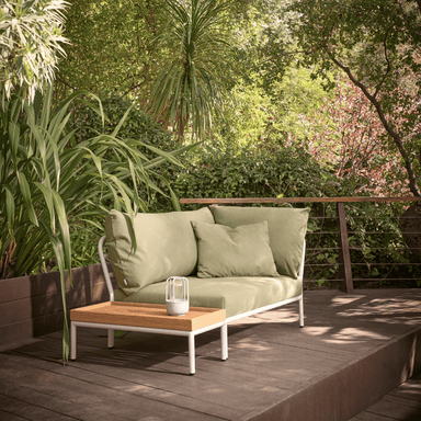 A light green right corner lounge sofa with white aluminum frame and a wooden side table set on a wooden deck patio surrounded by greenery