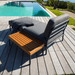 A wooden outdoor side table and dark gray outdoor lounge chair and dark gray outdoor ottoman set on a wooden deck poolside