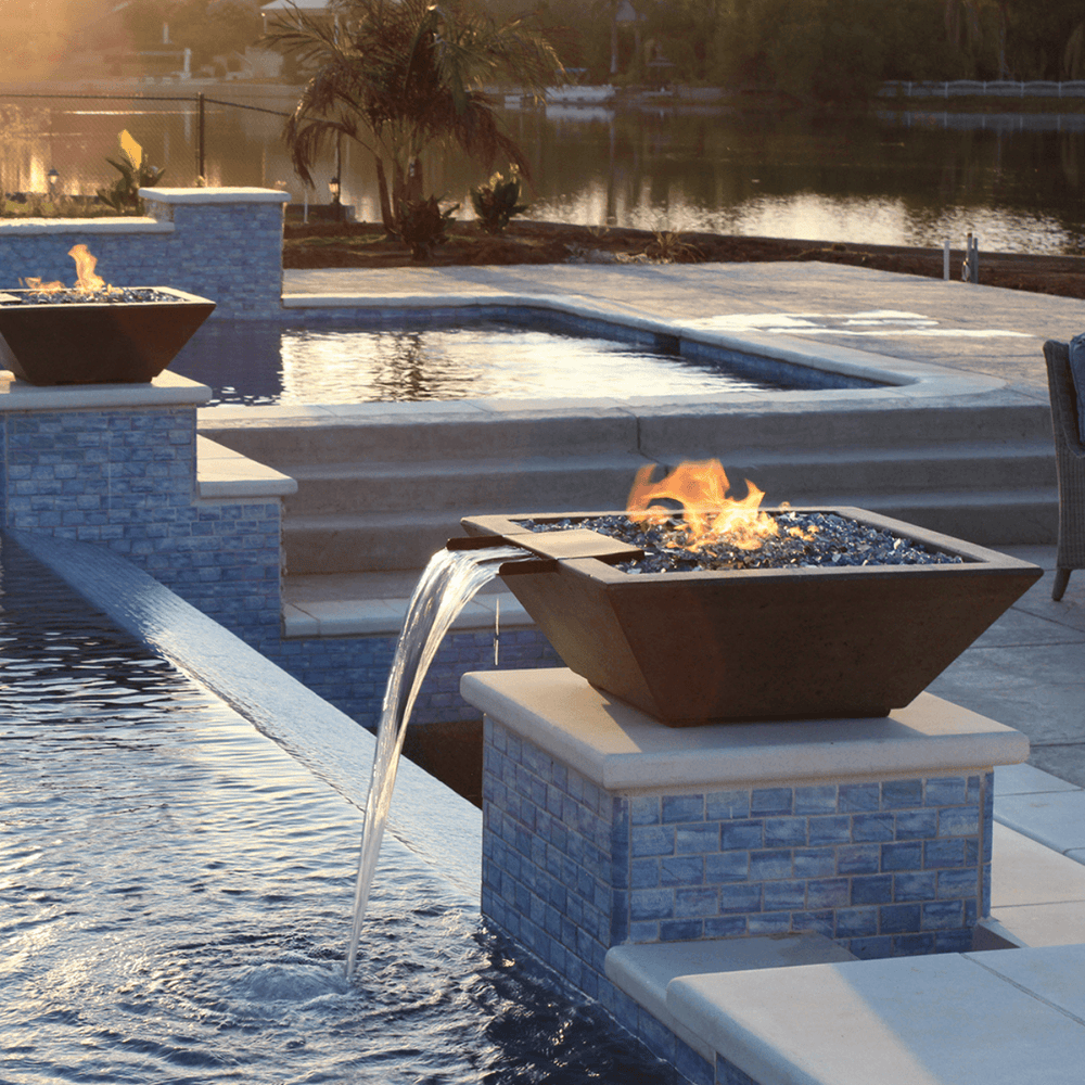 Two black tapered shaped square concrete fire water bowl with flowing water set on a raised platform at the corner of a pool