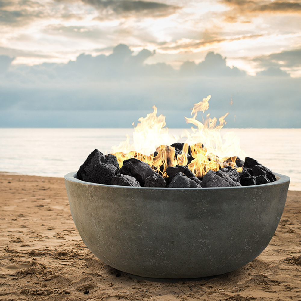 Grey round outdoor concrete fire bowl filled burning charcoal sits on a sandy beach