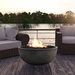 Grey round outdoor concrete fire bowl placed on a wooden deck outdoor surrounded by two wicker chairs with white cushion