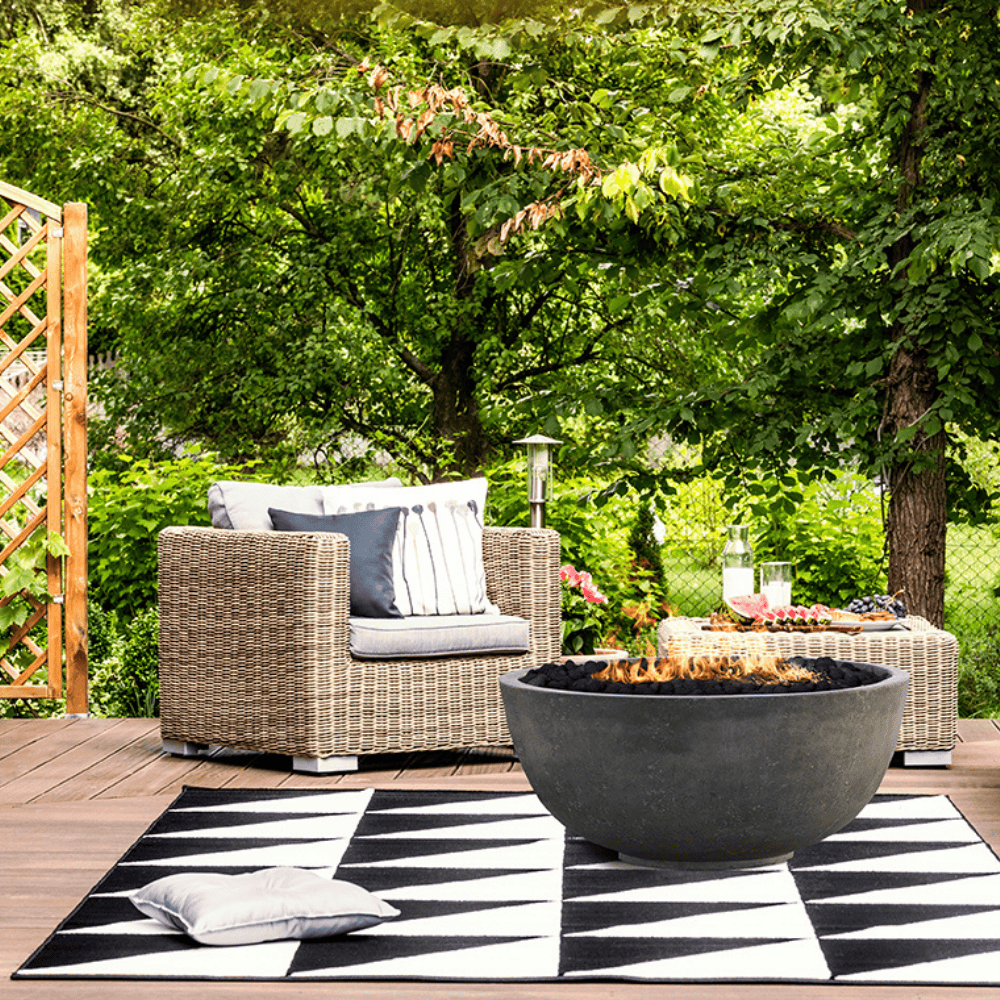 A grey round outdoor concrete fire bowl and wicker sofa placed on an outdoor patio setting surrounded by trees