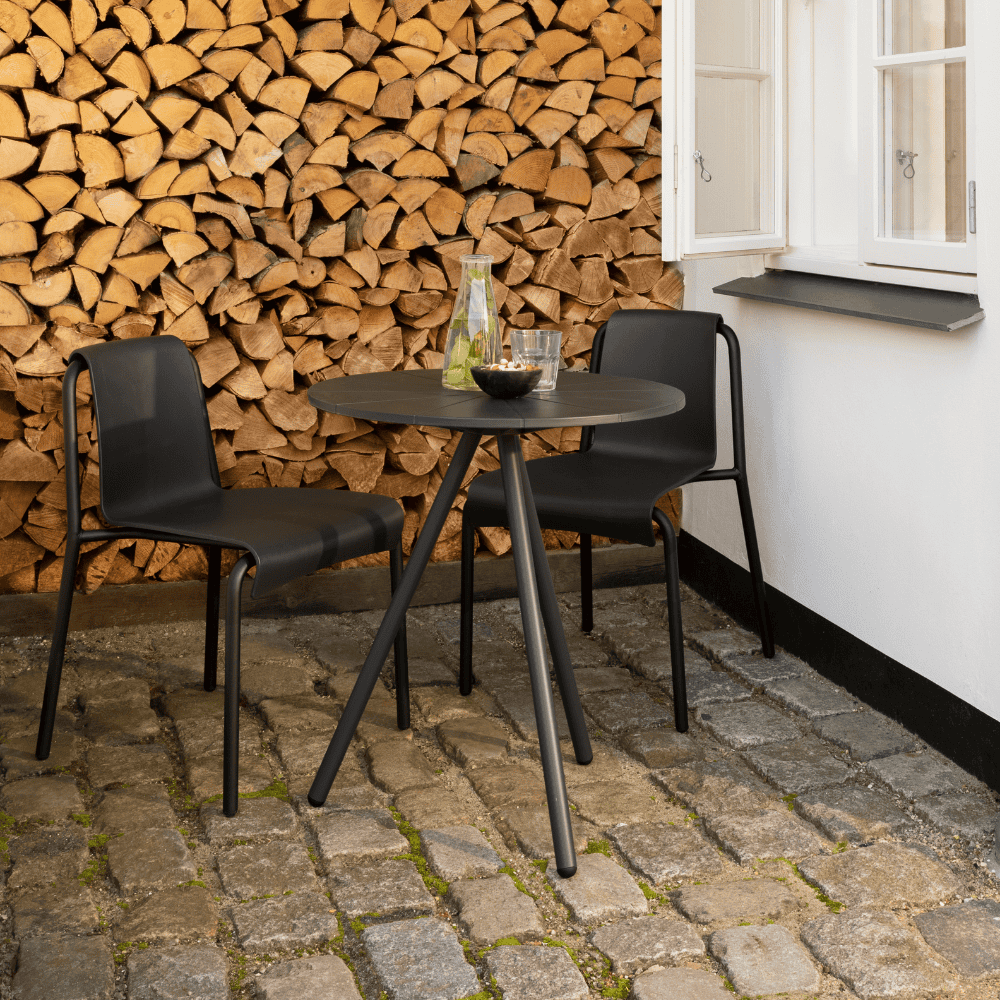 A black round outdoor coffee table and 2 black armless outdoor dining chairs placed on a cobblestone patio beside the window