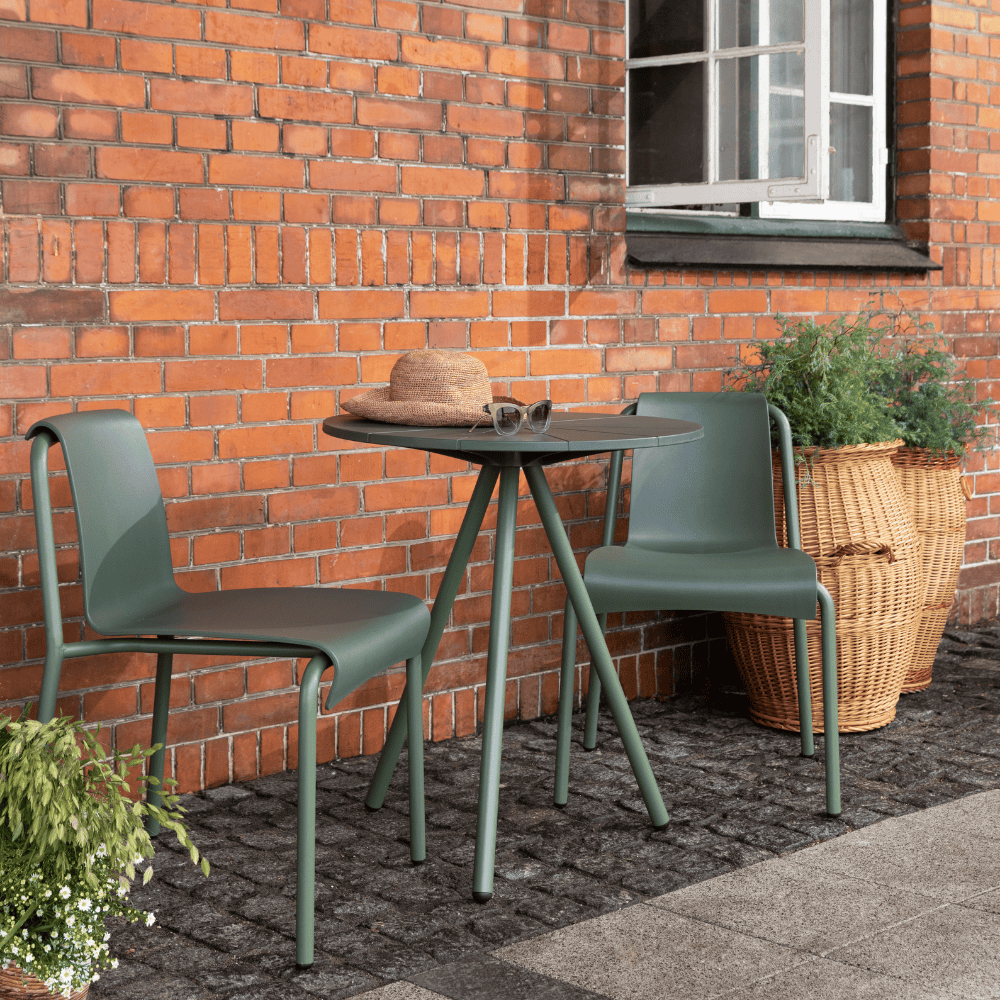 A green round outdoor coffee table and 2 green armless outdoor dining chairs set on a patio against orange brick wall