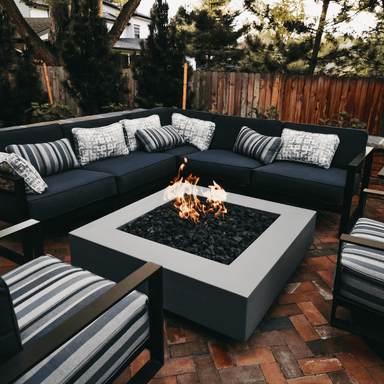 A grey square concrete fire pit table set in the center of an outdoor patio with dark blue sectional sofa and stripped chairs