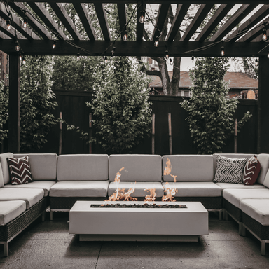 A light grey concrete rectangular fire pit table with sectional sofa sets on ab outdoor patio covered by a wooden pergola