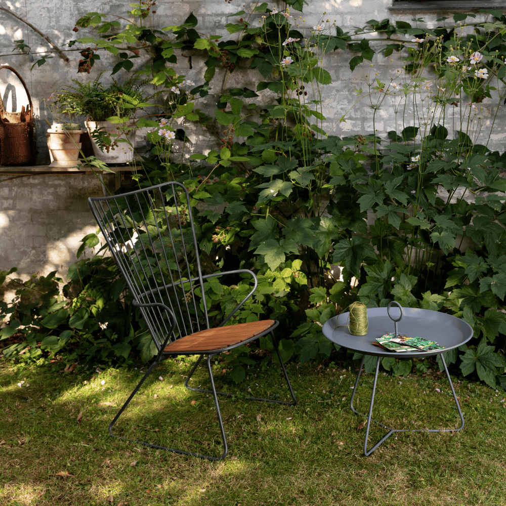 A black outdoor lounge chair with bamboo lamellas seat and a gray outdoor tray table set on a lawn area surrounded by plants