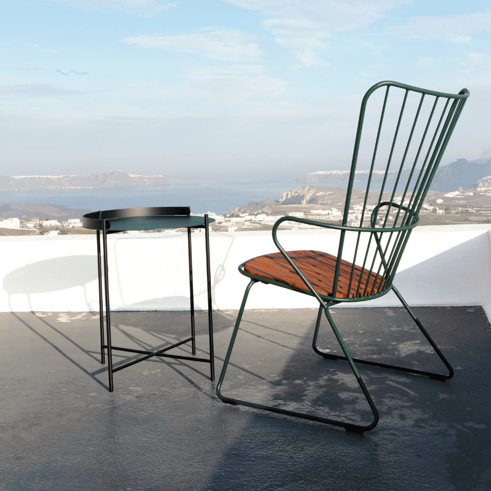 A dark green metal outdoor lounge chair with bamboo lamellas seat and a black outdoor tray table placed on a terrace