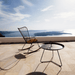 A black outdoor rocking chair with bamboo lamellas seat and an outdoor tray table set on a tiled patio with view of a sea and islands