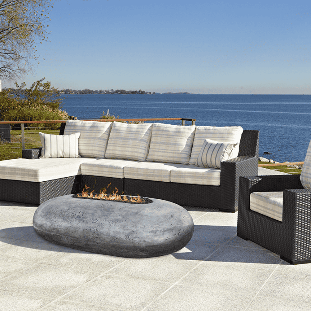 Grey pebble outdoor concrete oval fire table sets on an outdoor patio with modern sectional sofa and a view of a calm sea 