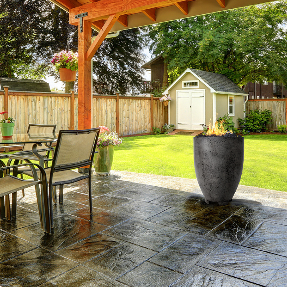 A grey outdoor concrete fire bowl with outdoor dining table and chairs placed on a tiled patio area with a wooden pergola