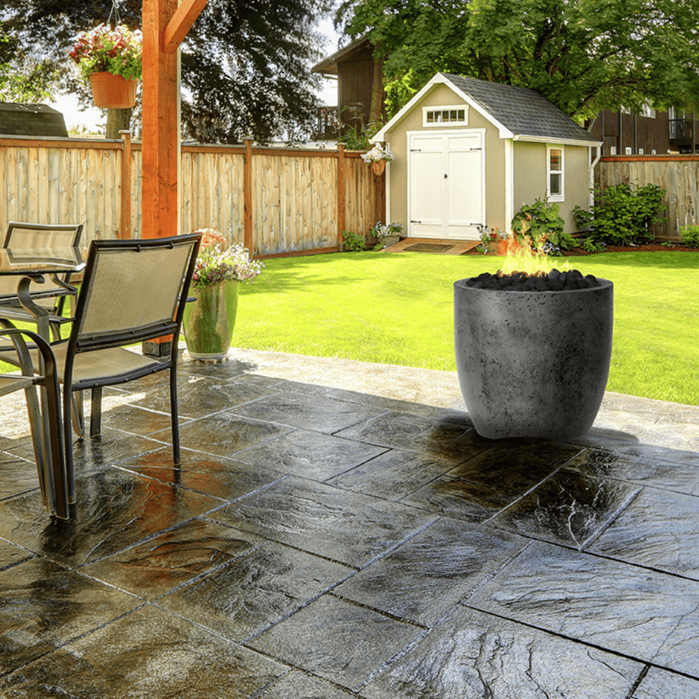 A grey round outdoor concrete fire bowl with outdoor dining table and chairs placed on a tiled patio area with a wooden pergola