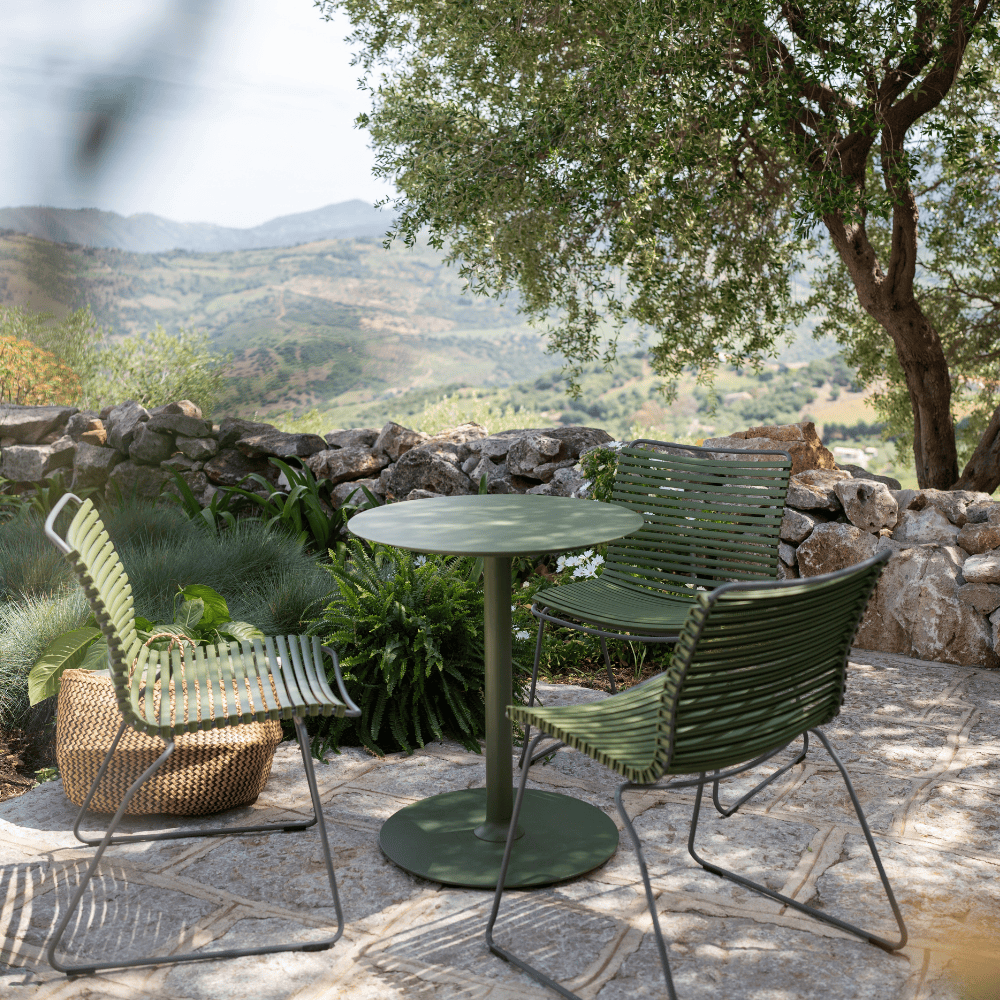 A green round outdoor coffee table and 3 green outdoor dining chairs set on a patio with stone wall surrounded by plants