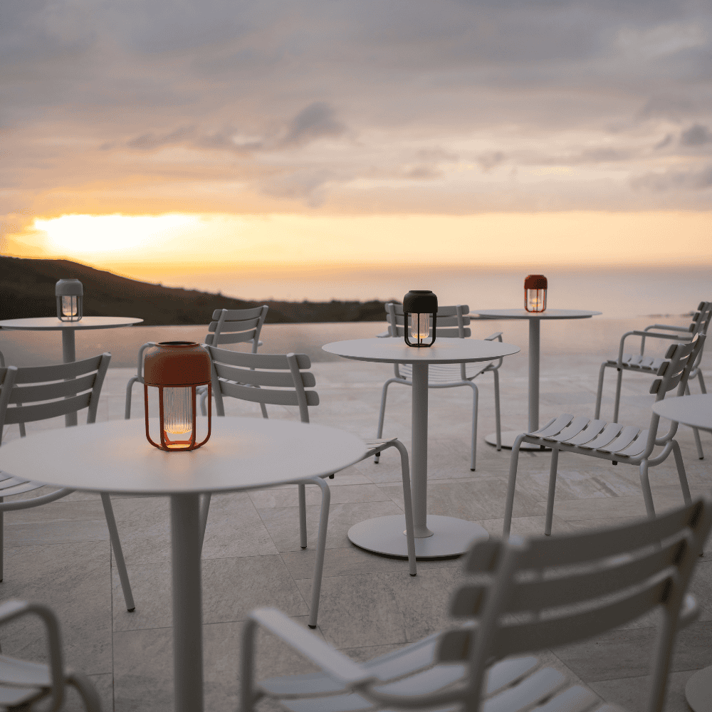 Several white round outdoor coffee tables and white outdoor dining chairs set on a tiled surface with view of a distant hill