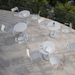 Several white round outdoor coffee tables and white outdoor dining chairs set on a tiled terrace with black railings