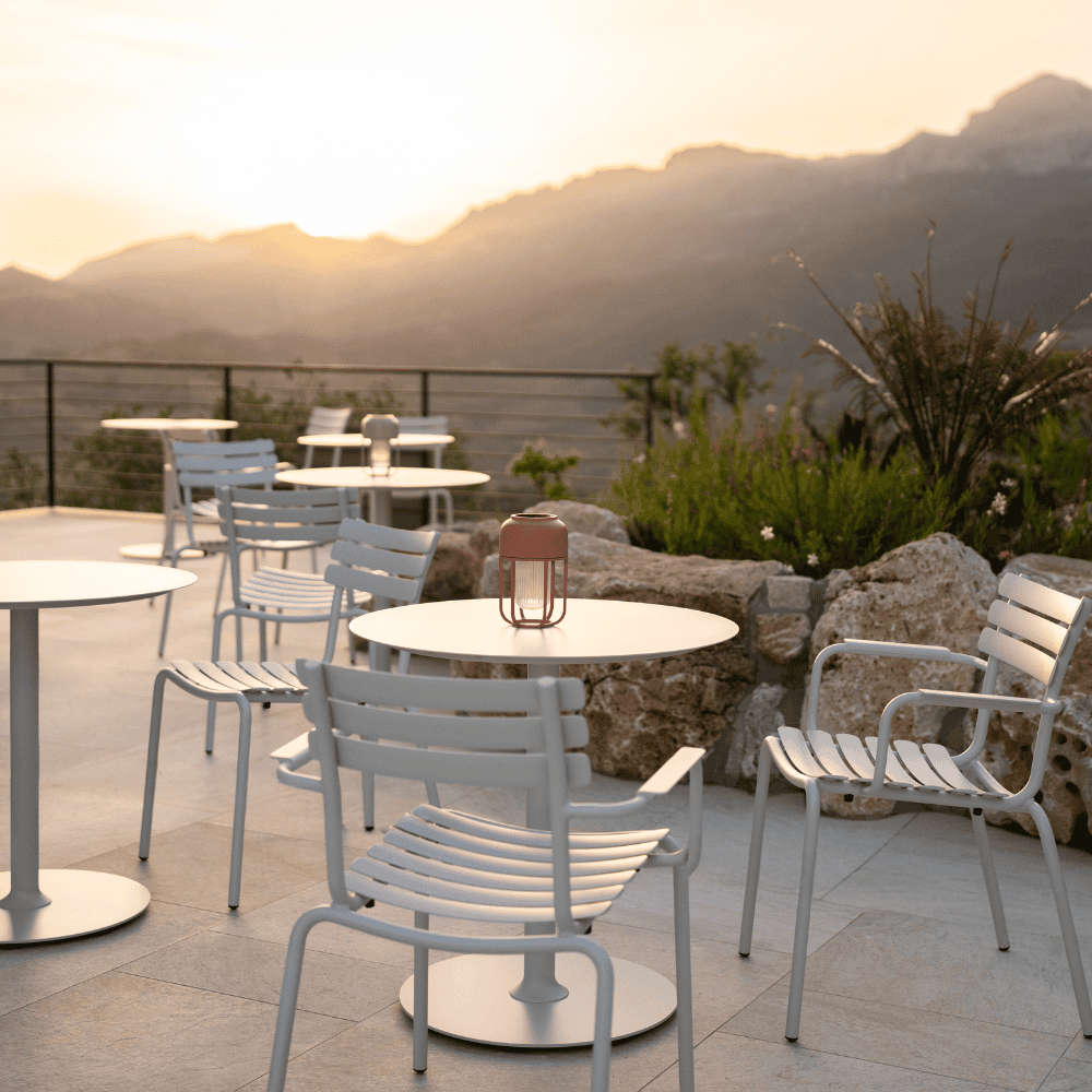 Several white round outdoor coffee tables and white outdoor dining chairs set on terrace with large stones and greenery