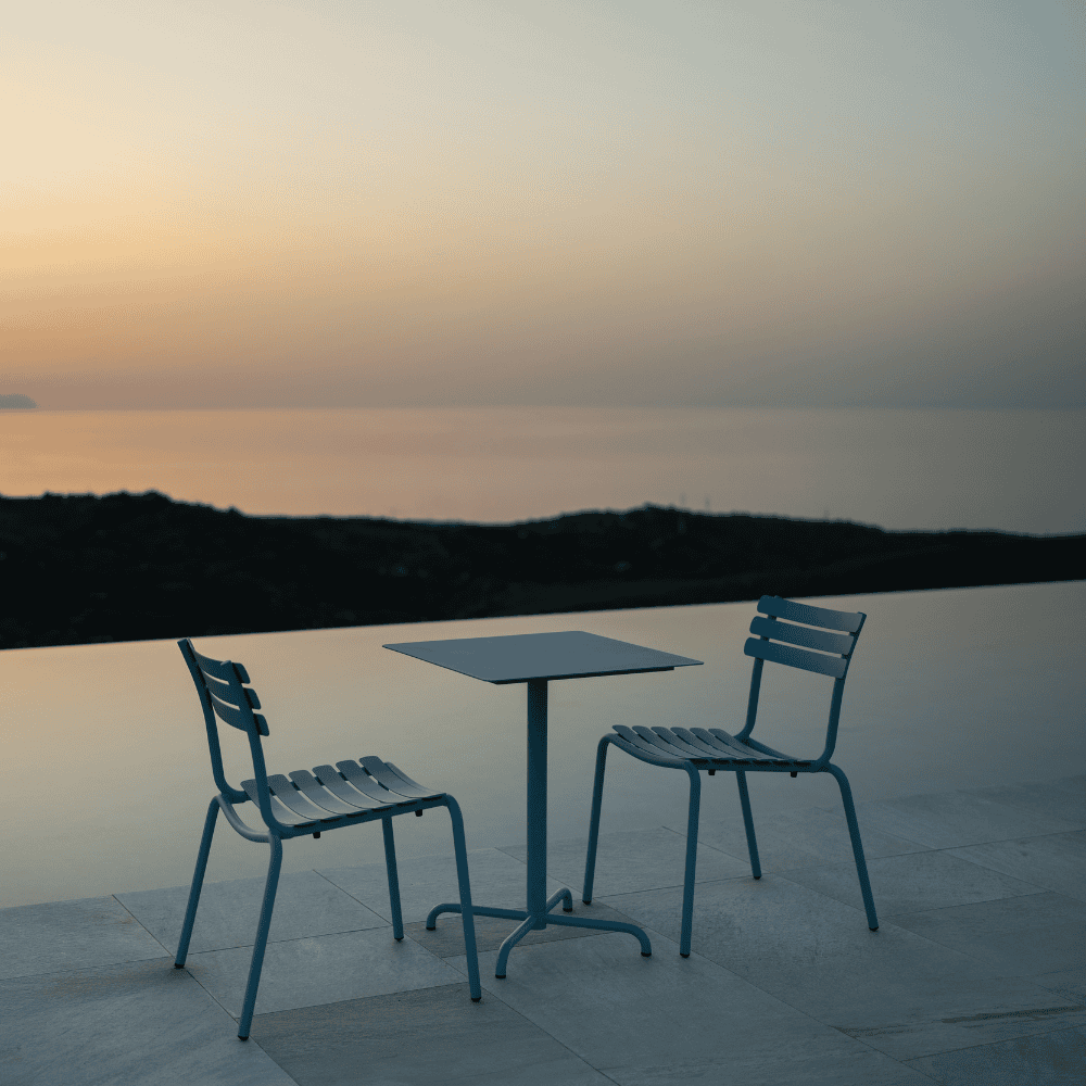 A blue square outdoor coffee table and 2 blue outdoor dining chairs placed on a tiled surface near the pool