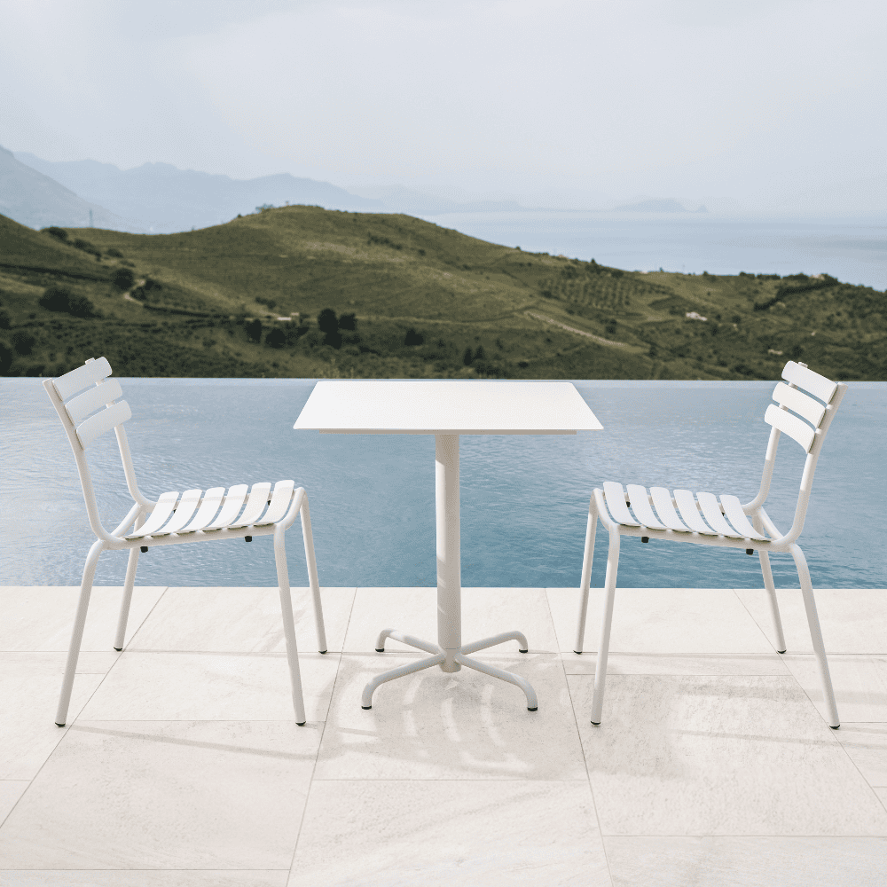 A white square outdoor coffee table and 2 white outdoor dining chairs placed beside the pool with view of a distant hills