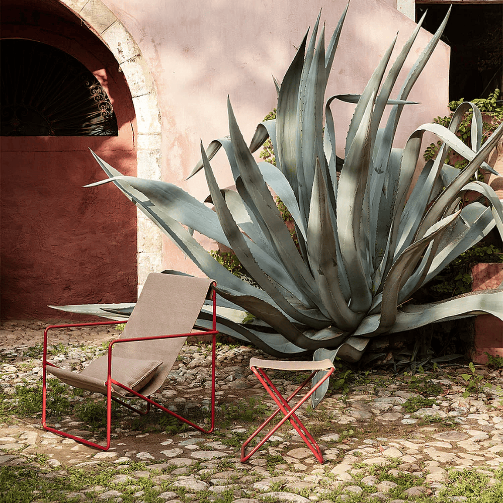modern light brown outdoor lounge chair with red steel frame placed on a rocky field beside large plant