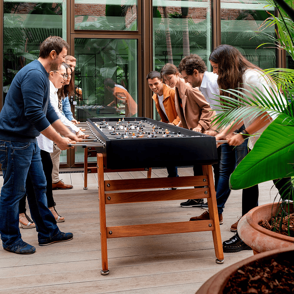 A group of people are playing a large black outdoor football table set on a wooden deck patio in front of a glass door