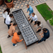 A group of people are actively playing a large black outdoor football table situated outdoors on a wooden deck