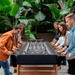 A group of people are playing a large black outdoor football table in an outdoor area surrounded by lush green plants