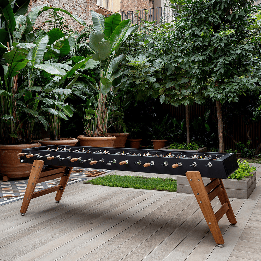 A large black outdoor football table with wooden legs set on a wooden deck surrounded by green large plants in a large pots