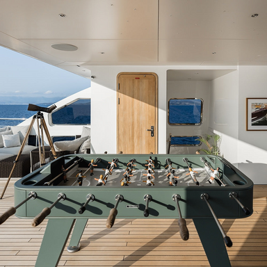 A green outdoor football table with wooden handles is set on a deck of a luxurious yacht in front of a wooden door