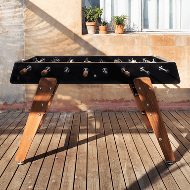 A black outdoor football table with wooden legs and handles set on a wooden deck in front of the wall with a window and potted plants