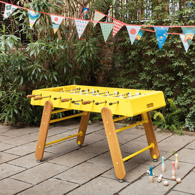 A yellow outdoor football table with wooden legs and handles set up outdoors on a stone patio surrounded by lush green plants