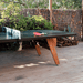 A stationary black outdoor ping pong table with wooden legs set on a wooden deck surrounded by plants and wooden fence