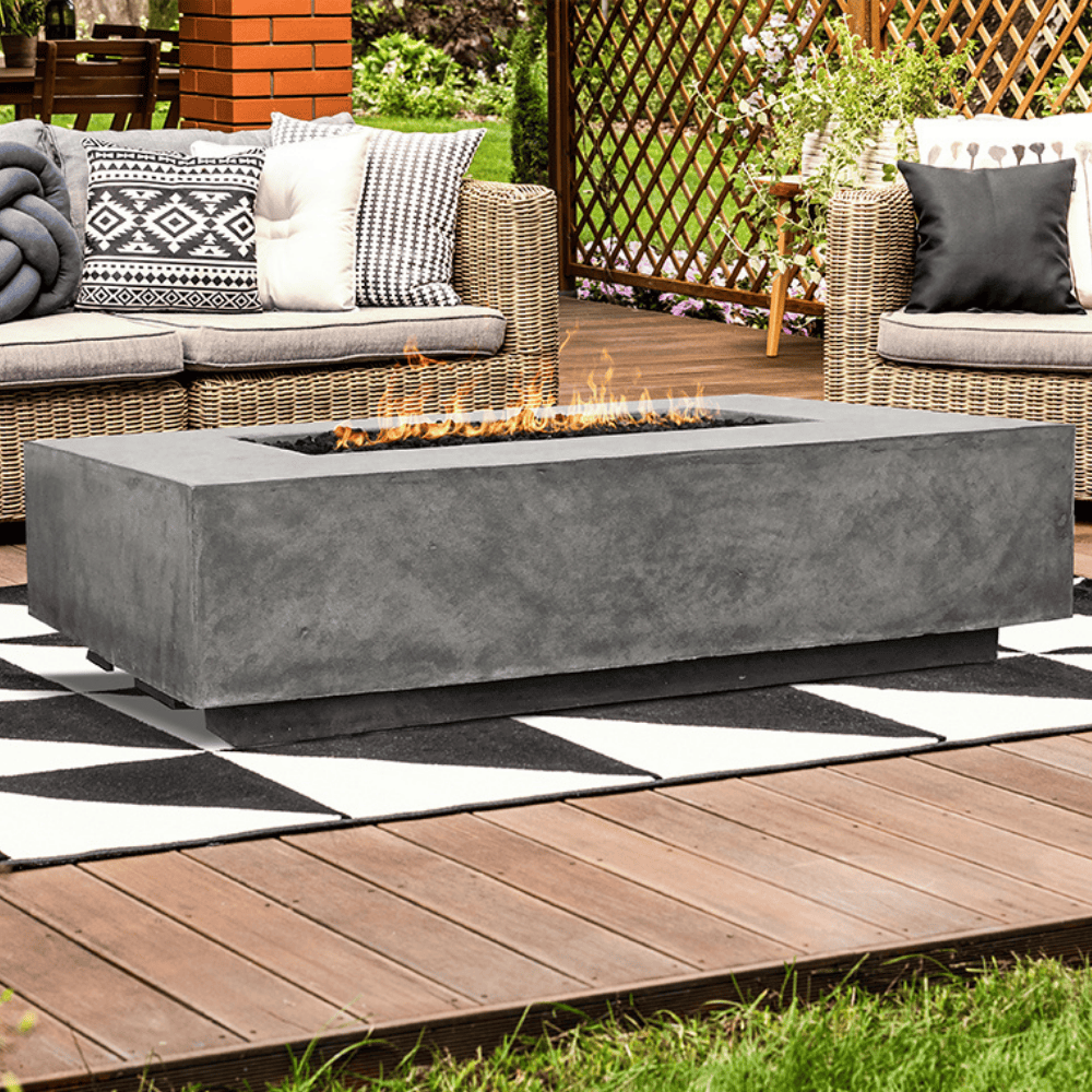 A grey outdoor concrete rectangular fire pit table set on a wooden deck patio beside wicker sofa with cushion