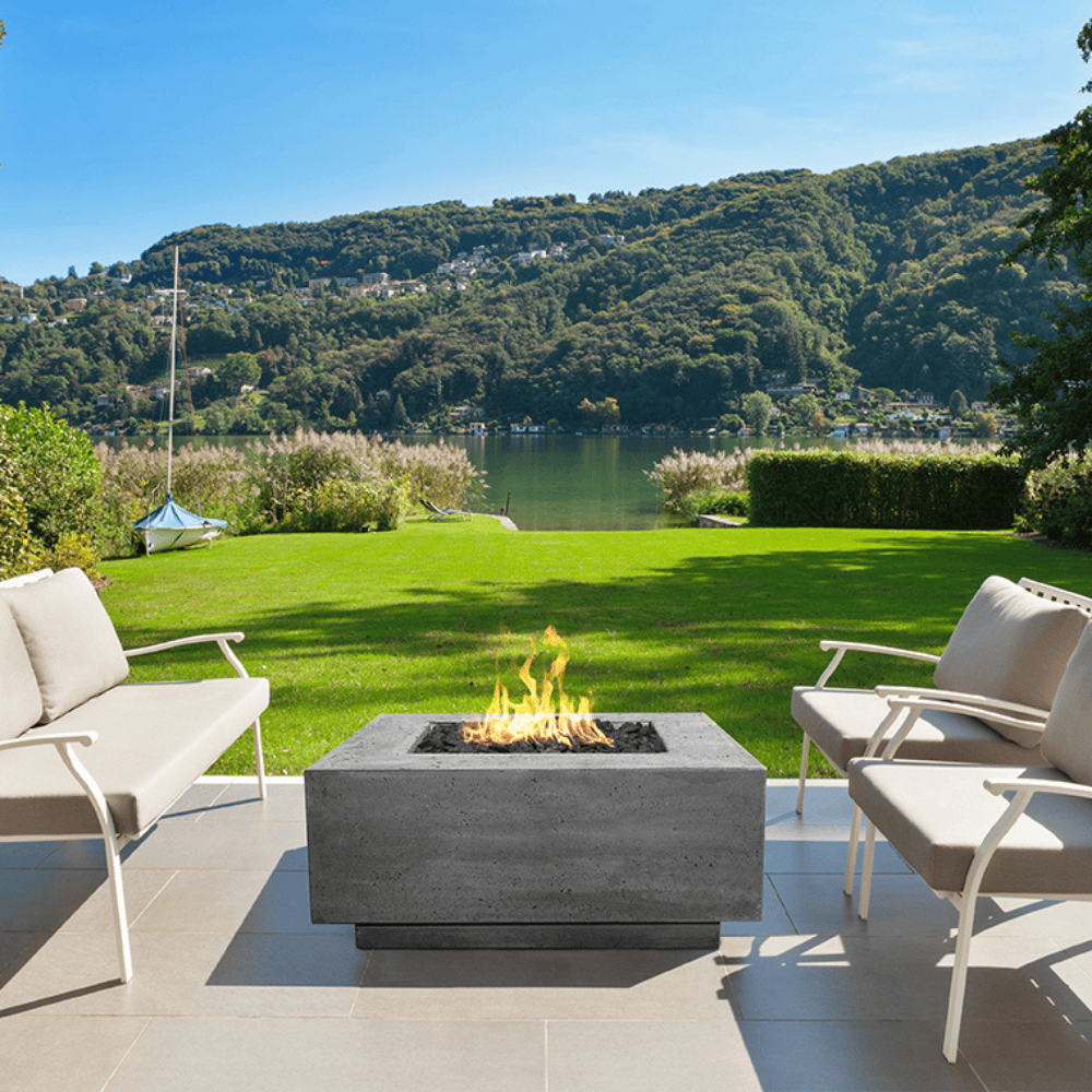 A grey square outdoor concrete fire table with sofa and chairs set on a tiled surface in an outdoor patio with green lawn