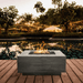 A grey square outdoor concrete fire table on a wooden deck beside the pool with plants and trees around with light strings