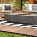 A grey concrete outdoor rectangular fire pit table with wicker sofa and chair with cushion set on a wooden deck patio garden