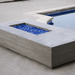 A grey concrete outdoor rectangular fire pit table with blue fire glass placed beside the pool