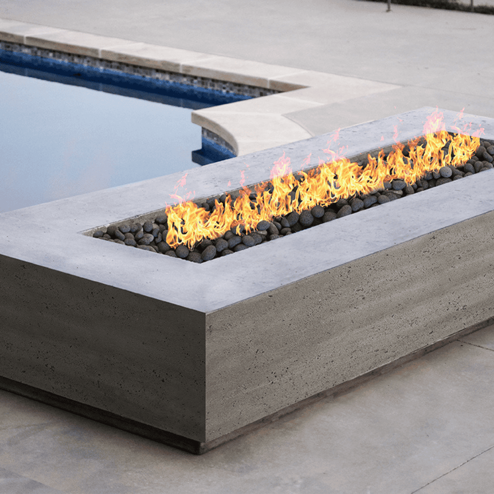 A grey concrete outdoor rectangular fire pit table with flames burning over lava rocks placed beside the pool