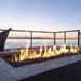 Close up view of a concrete outdoor rectangular fire pit table with flames with a view of a calm sea and distant mountains