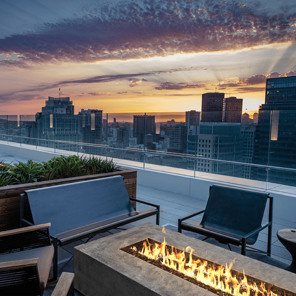 A concrete outdoor rectangular fire pit set on a patio accompanied by black metal sofa and chair with a city view background