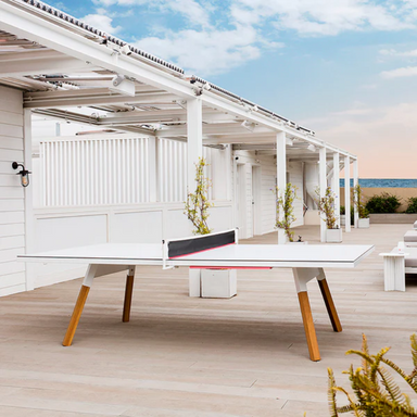 A white outdoor ping pong table with wooden legs set on a wooden deck patio with several potted plants