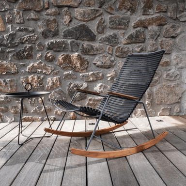 A black outdoor rocking chair with bamboo armrest and black round outdoor tray table placed on a wooden deck with stone wall