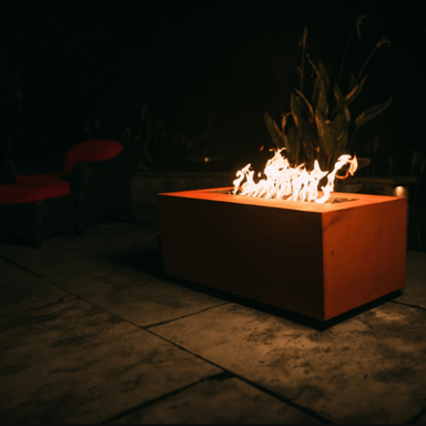 Orange rectangular powder coated metal fire pit with vibrant flames on an outdoor patio at night accompanied by plants and two red chairs